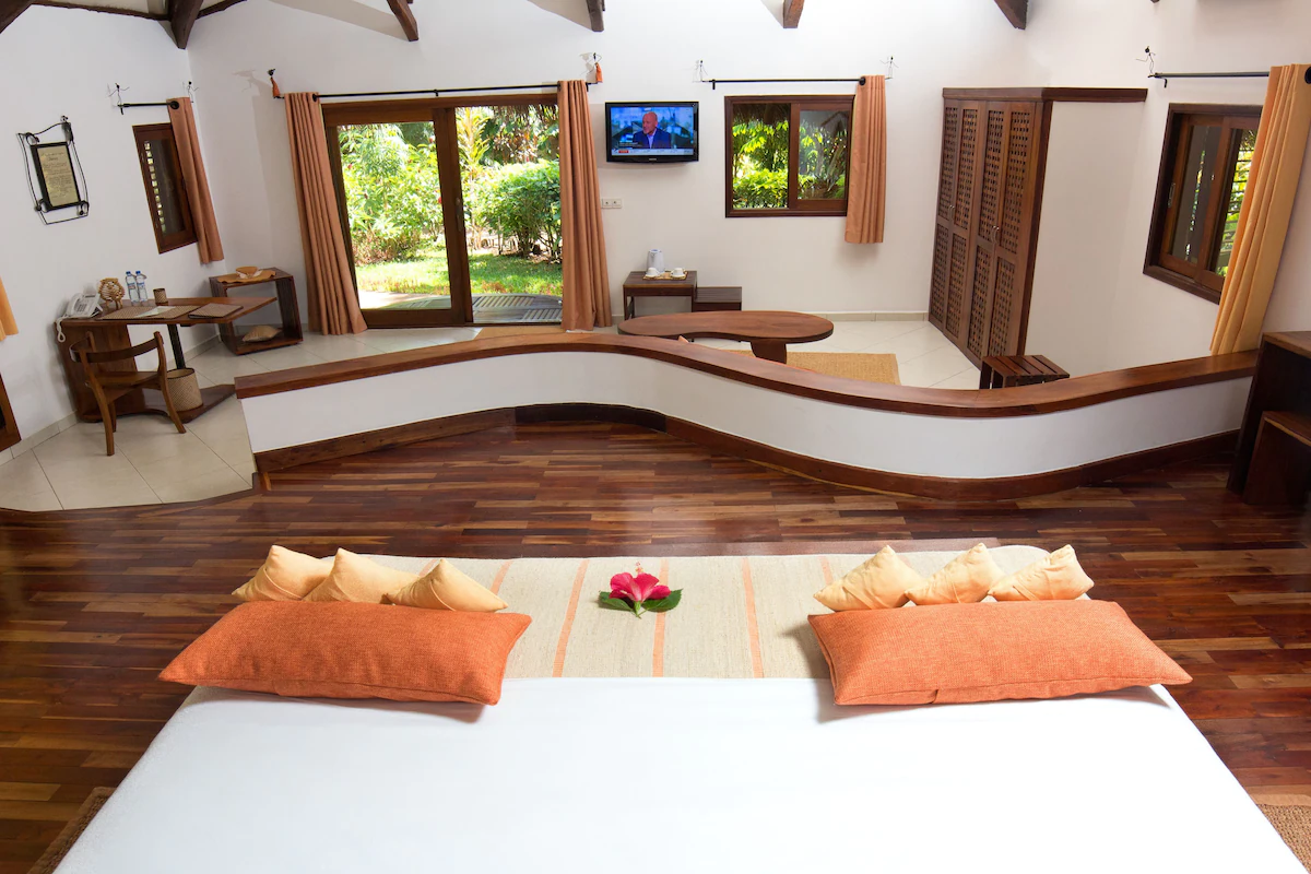 Bedroom with large bed, orange pillows, wooden floor, white walls, and open window showing greenery.