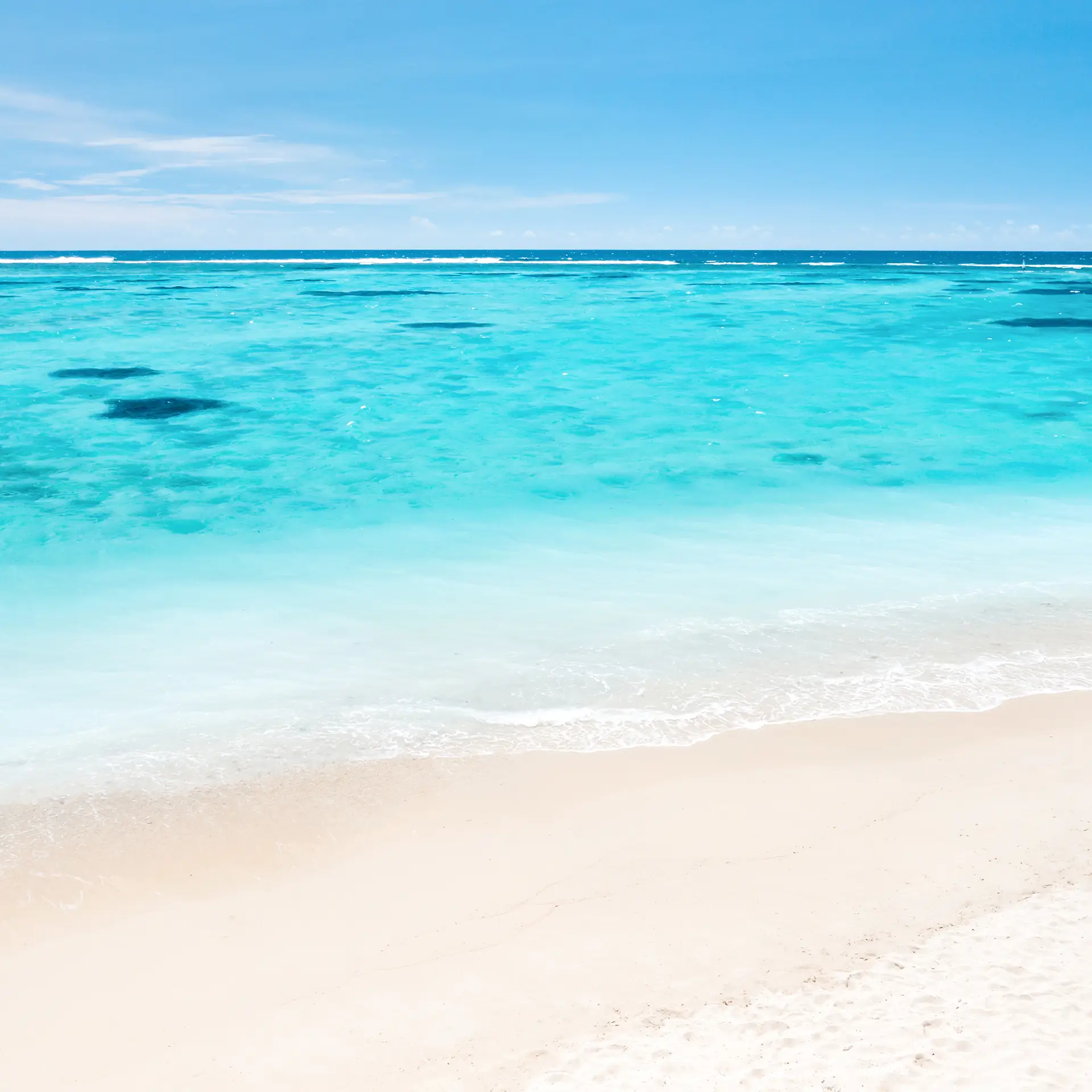 Clear turquoise ocean water gently waves onto a white sandy beach under a blue sky.